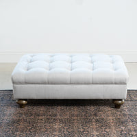 The Chesterfield Footstool in Warm Grey, featuring a tufted design and short wooden legs, sits on a patterned rug against a plain white wall.