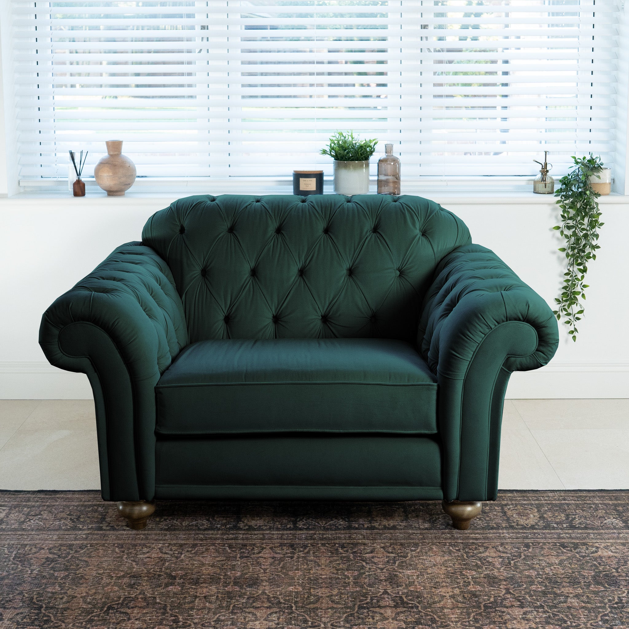 A Chesterfield Velvet Loveseat in Bottle Green, handcrafted in England, enhances a bright room with white blinds, lush plants, and a decorative rug.