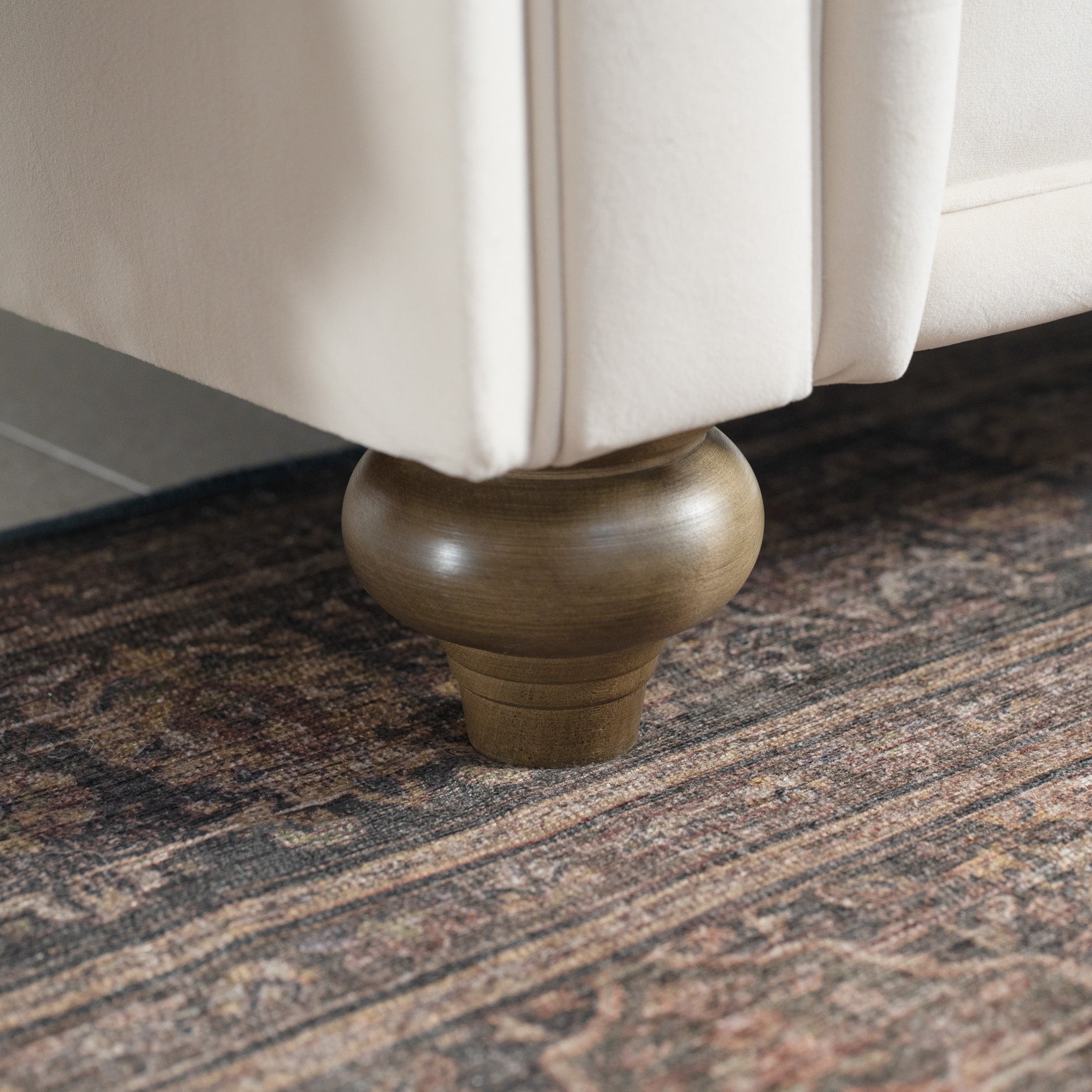 Close-up of a wooden sofa leg on a patterned carpet, highlighting the craftsmanship of sustainable birch and beech, reminiscent of the Chesterfield Velvet Loveseat in Cream handcrafted in England.