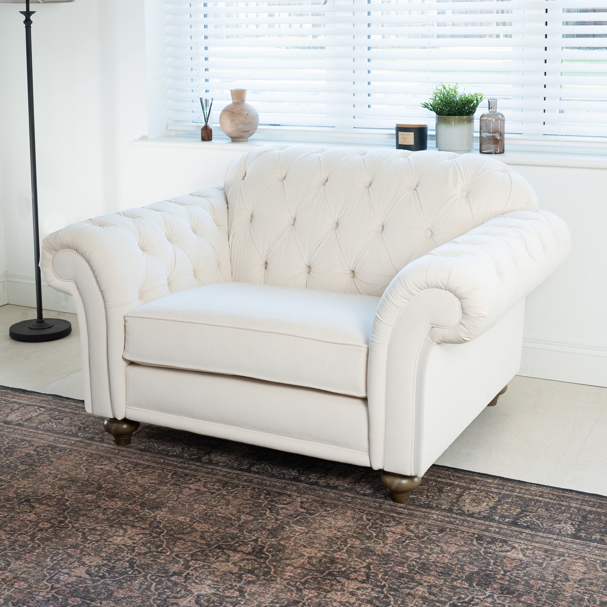 A white tufted armchair on a patterned rug sits in a bright, minimalist room with a window, next to the elegant Chesterfield Velvet Loveseat in Cream, handcrafted in England.