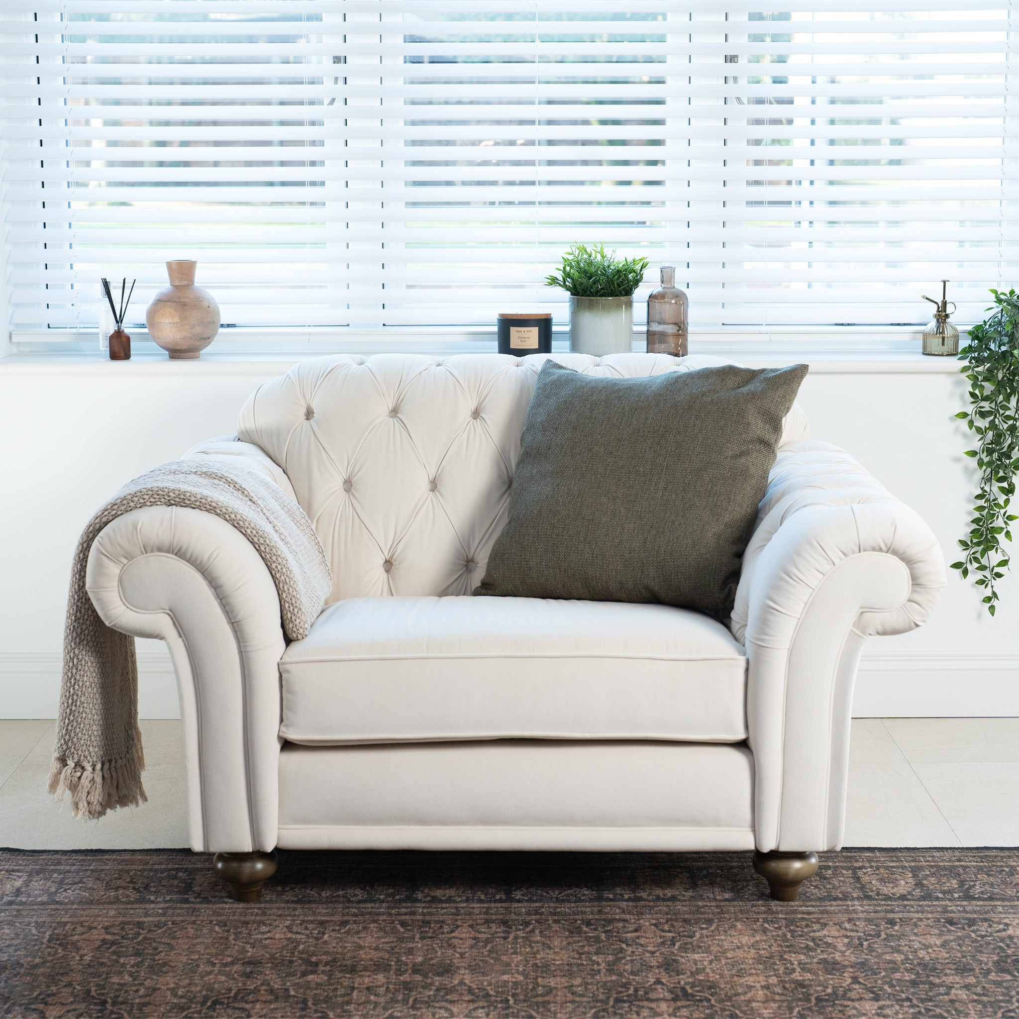 The Chesterfield Velvet Loveseat in Cream, accented with a gray pillow and blanket, handcrafted in England, sits in front of window blinds, flanked by plants and decor.