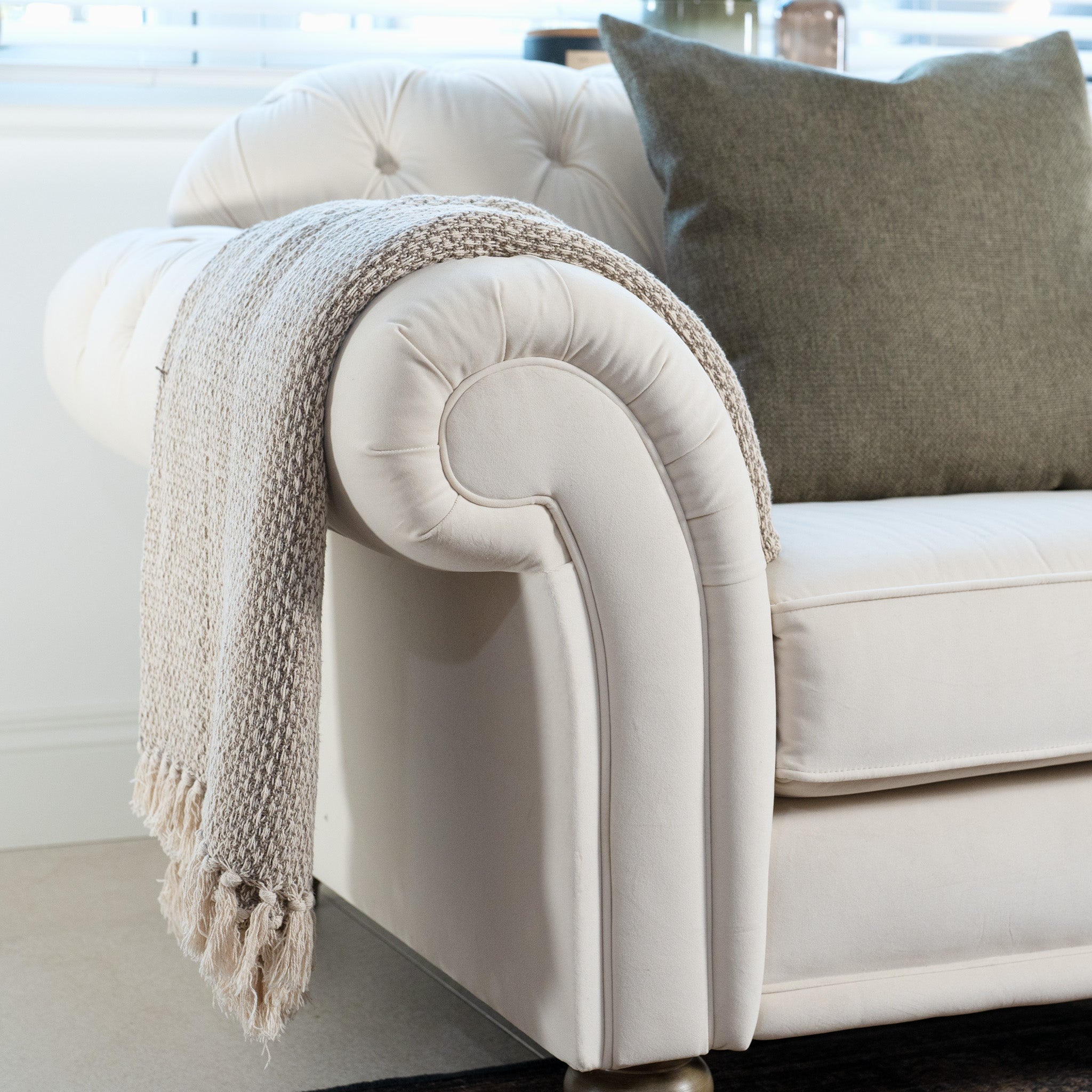 The handcrafted Chesterfield Velvet Loveseat in Cream, made in England, is adorned with a gray pillow and a textured beige throw draped over its armrest in a bright room.