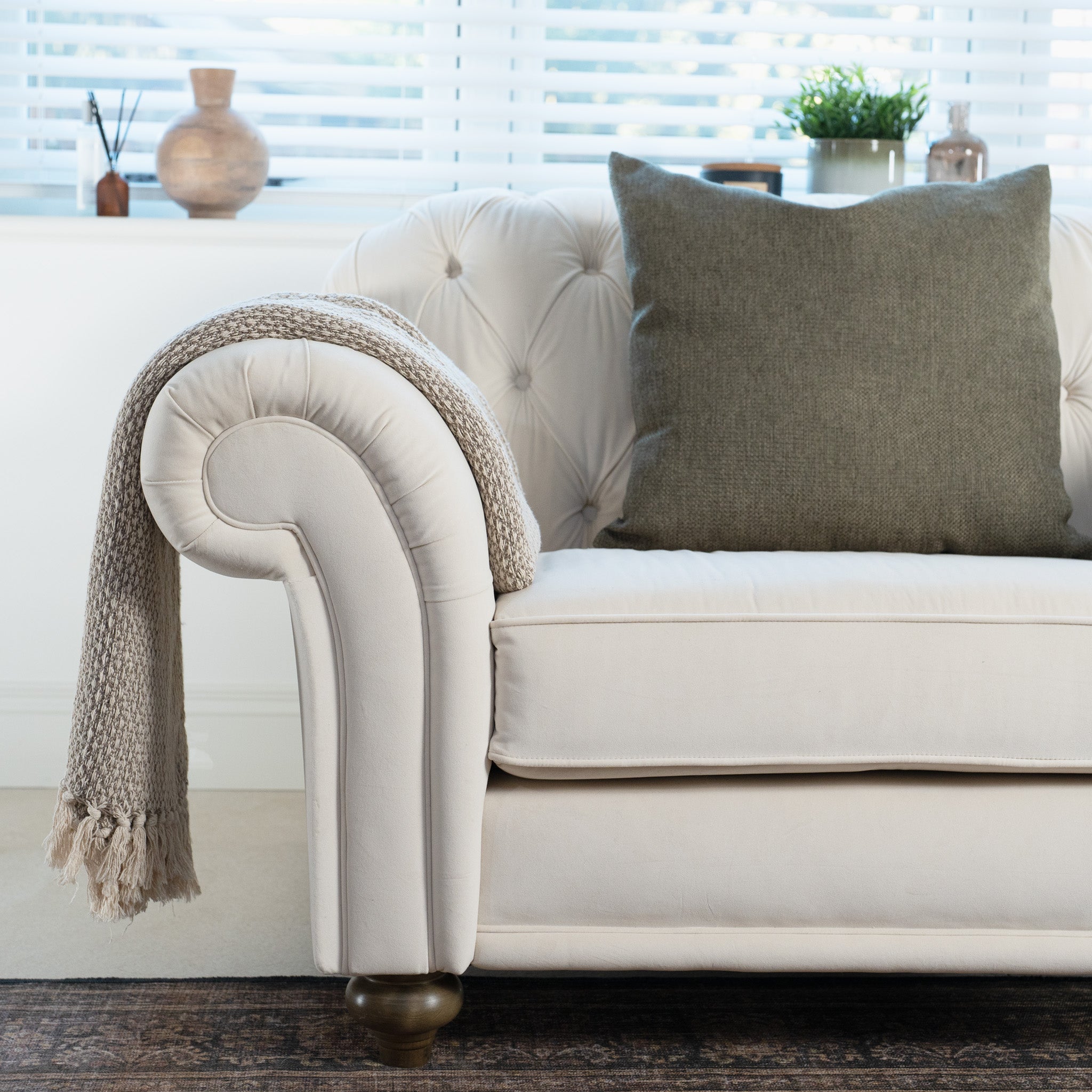 A cozy Chesterfield Velvet Loveseat in Cream, handcrafted from sustainable birch and beech in England, is adorned with a green pillow and beige throw as sunlight filters through nearby window blinds.