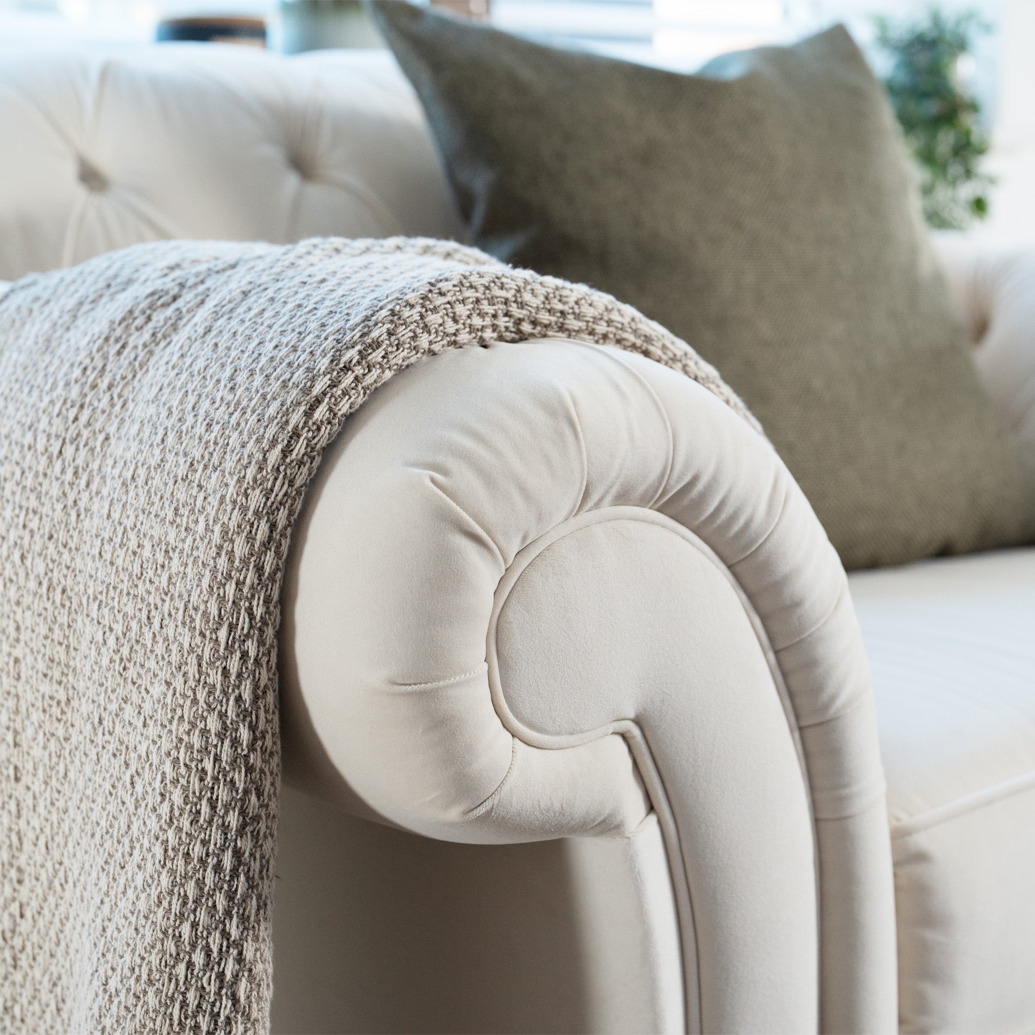 The Chesterfield Velvet Loveseat in cream, handmade in England from sustainable birch and beech, showcases a textured blanket on the armrest, complemented by a green cushion behind.