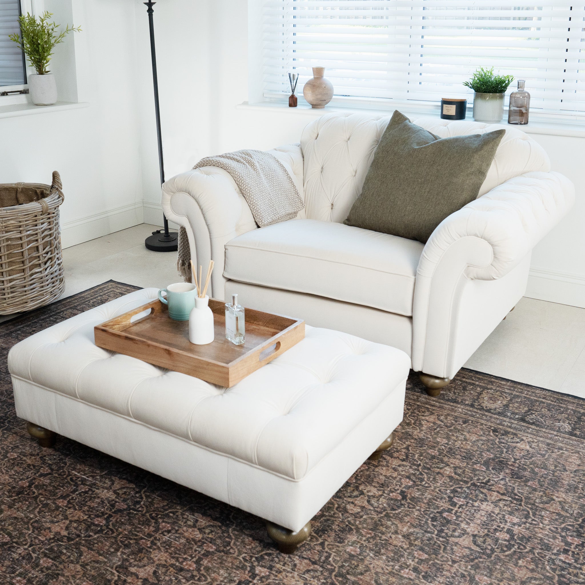A cozy living room features a white armchair, ottoman, and wooden tray on a patterned rug, exuding classic elegance. The Chesterfield Velvet Loveseat in Cream, handcrafted in England, adds sophistication to the space with its timeless style.