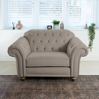 A plush beige armchair with a tufted backrest sits in a well-lit room, perfectly complementing the Chesterfield Velvet Loveseat in Mink. Plants and vases on the windowsill enhance the inviting and elegant atmosphere.