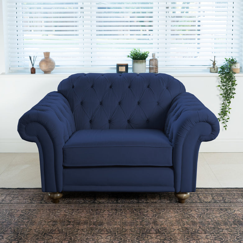 The Chesterfield Velvet Loveseat in Royal Blue graces the bright room with white blinds, its elegance accentuated by decor on the windowsill. Handmade in the UK, this piece adds sophistication to any setting.