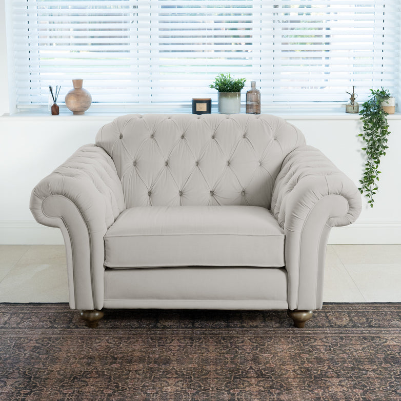 The Chesterfield Velvet Loveseat in Feather Grey, with its tufted elegance, sits by the window surrounded by blinds and potted plants, exuding timeless charm.