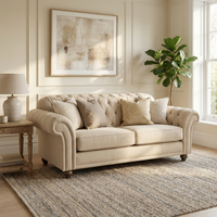 The Chesterfield 2 Seat Sofa in Beige with pillows sits in a bright living room, accented by a rug, side table, lamp, artwork, and potted plant—handcrafted in England for timeless elegance and classic style.