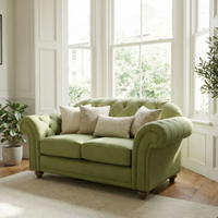 The Chesterfield 2 Seat Sofa in Olive, adorned with assorted cushions, showcases classic British craftsmanship in a sunlit living room filled with large windows and vibrant greenery.