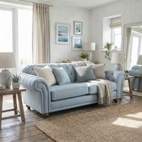 The Chesterfield 2 Seat Sofa in Sky Blue adds timeless elegance with neutral pillows, surrounded by plants, lamps, and framed art in a bright, cozy living room; handcrafted in England.