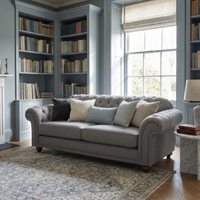 The Chesterfield 2 Seat Sofa in Smoke Grey sits in a cozy library with assorted cushions, bookshelves, a large window, and a patterned rug—elegantly handcrafted in England.