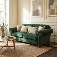 The Chesterfield 2 Seat Velvet Sofa in Bottle Green with beige pillows sits in a bright living room, paired with chic decor, a coffee table, and a floor lamp.