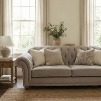A Chesterfield 2 Seat Velvet Sofa in Mink with throw pillows is placed in a bright living room featuring large windows, curtains, and a side table.