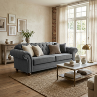 A cozy living room featuring the Chesterfield 2 Seat Velvet Sofa in Steel Grey, a coffee table, wall art, and sunlight streaming through large windows.