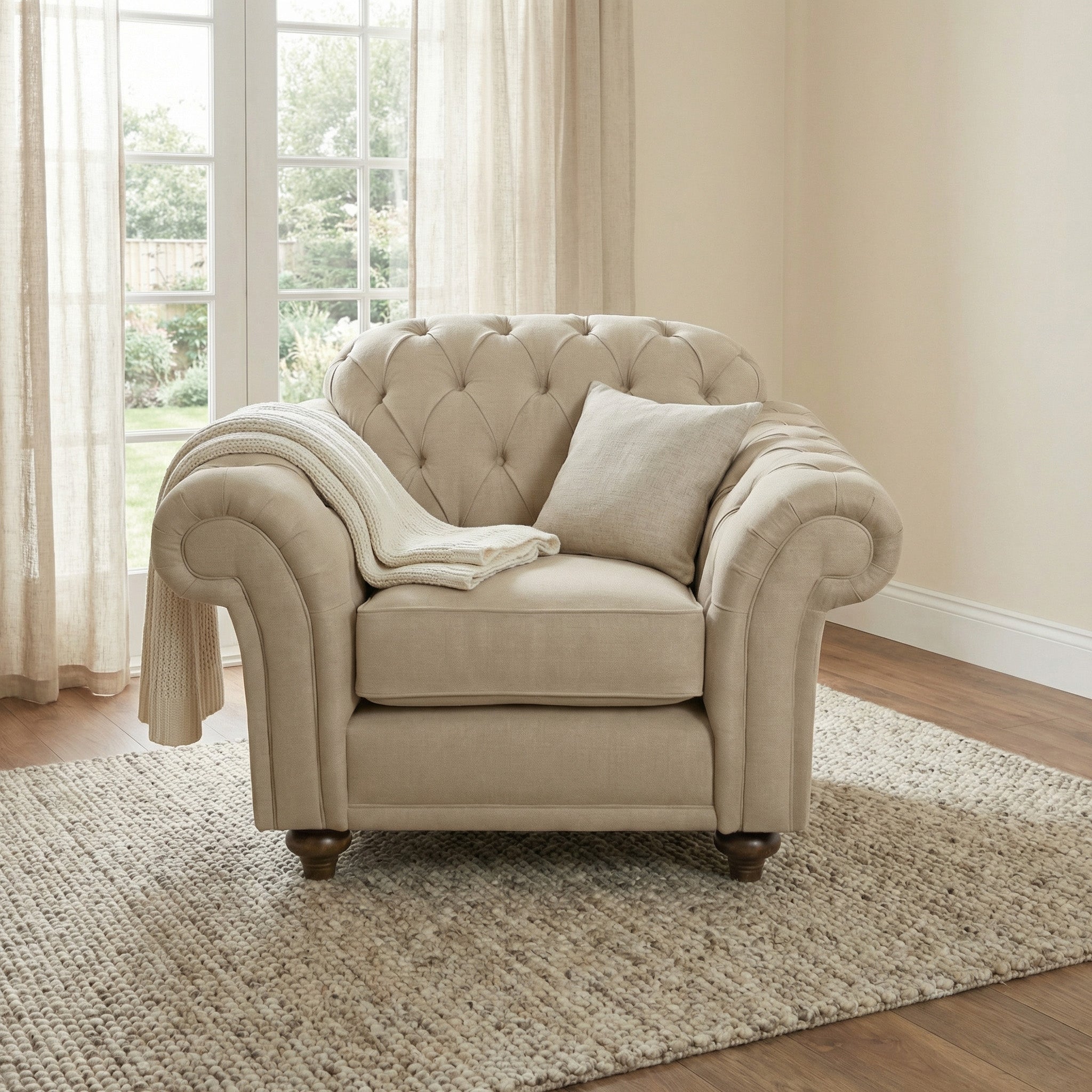 The Chesterfield Armchair in Beige sits on a rug in a sunlit room with large windows, accessorized with a pillow and throw blanket—an inviting piece that highlights traditional British craftsmanship.