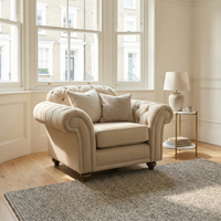 The Chesterfield Armchair in Beige, handcrafted in England, is placed by large windows in a bright, cozy living room with pillows, a rug, and a side table.
