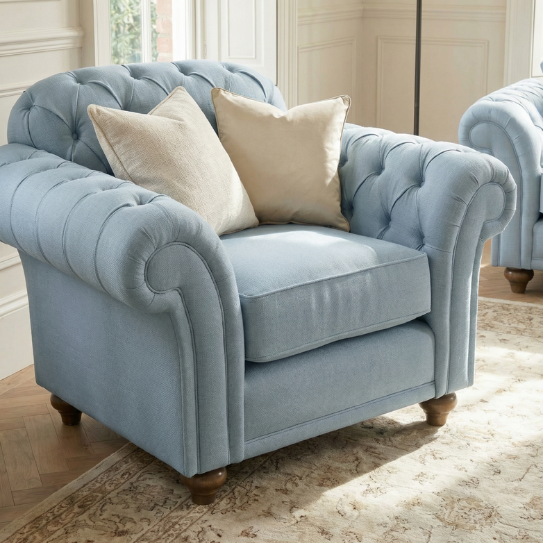 The Chesterfield Armchair in Sky Blue, featuring two beige pillows, is placed on a patterned rug in a bright room.
