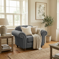 A cozy living room featuring a Chesterfield Armchair in Smoke Grey, complemented by cream pillows, a knit throw, a side table, and a potted plant.