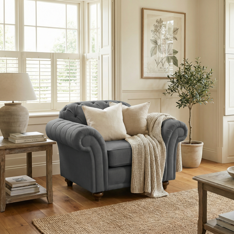 A cozy living room featuring a Chesterfield Armchair in Smoke Grey, complemented by cream pillows, a knit throw, a side table, and a potted plant.