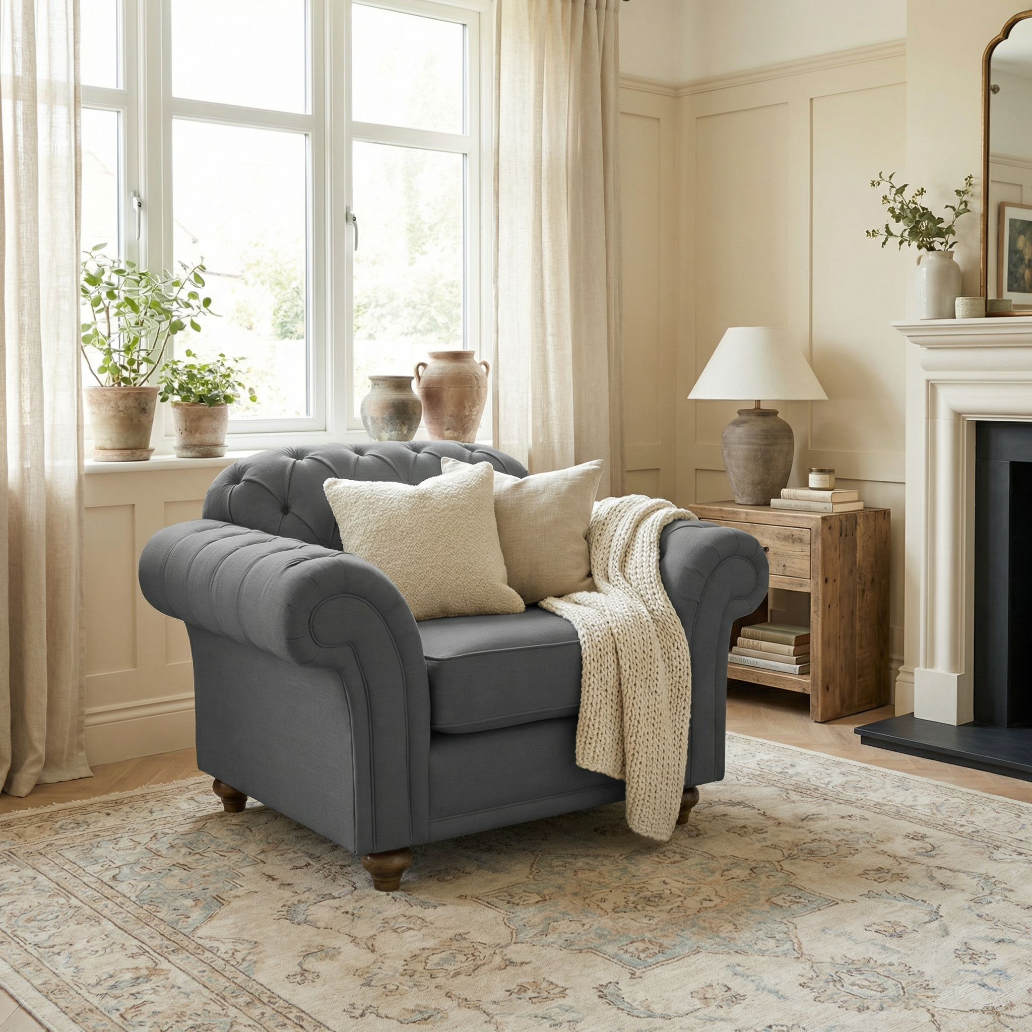 The Chesterfield Armchair in Smoke Grey, adorned with cushions and a blanket, sits in a sunlit living room filled with plants and neutral decor.