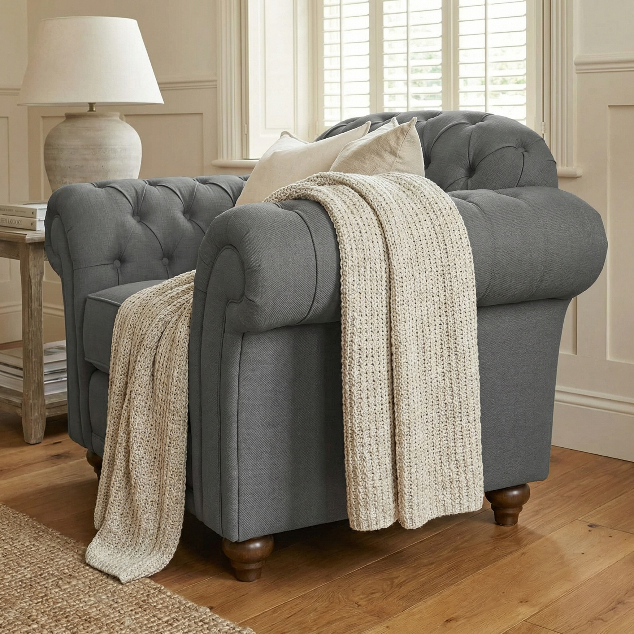 A Chesterfield Armchair in Smoke Grey, adorned with a cream knit throw and cushion, sits by the window in a cozy room, reflecting traditional British design.