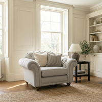 A Chesterfield Armchair in Warm Grey with cushions sits by a bright window in a stylish, neutral-toned living room, highlighting the elegance of handcrafted British furniture.