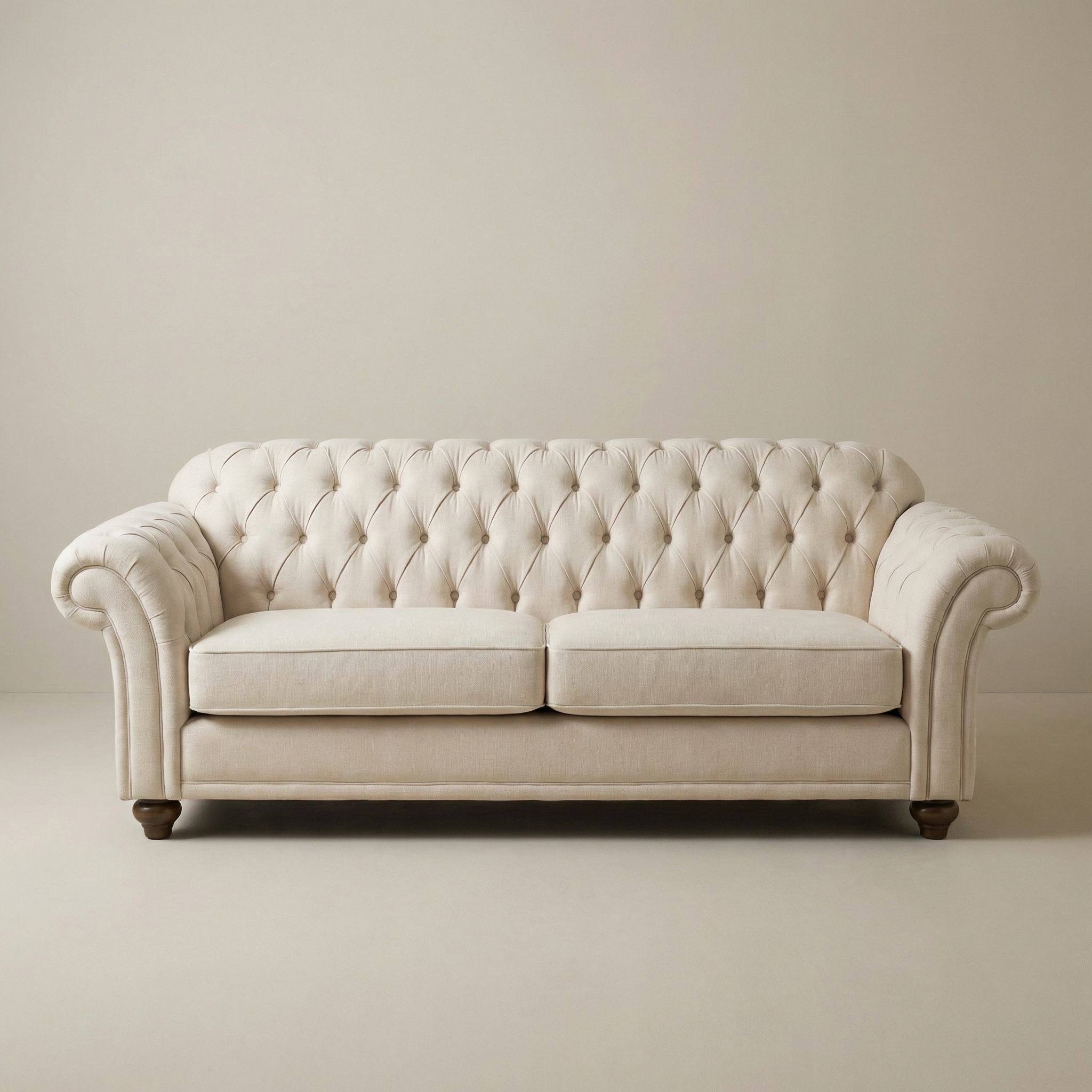 The Chesterfield 3 Seat Sofa in Ivory, featuring tufted upholstery, rolled arms, and wooden legs made from sustainably sourced wood, sits against a plain, light-colored wall and floor.