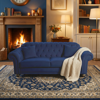 The Chesterfield 3 Seat Velvet Sofa in Royal Blue, paired with a throw blanket, brings warmth and invites comfort to the cozy living room alongside a glowing fireplace and elegant lamps.