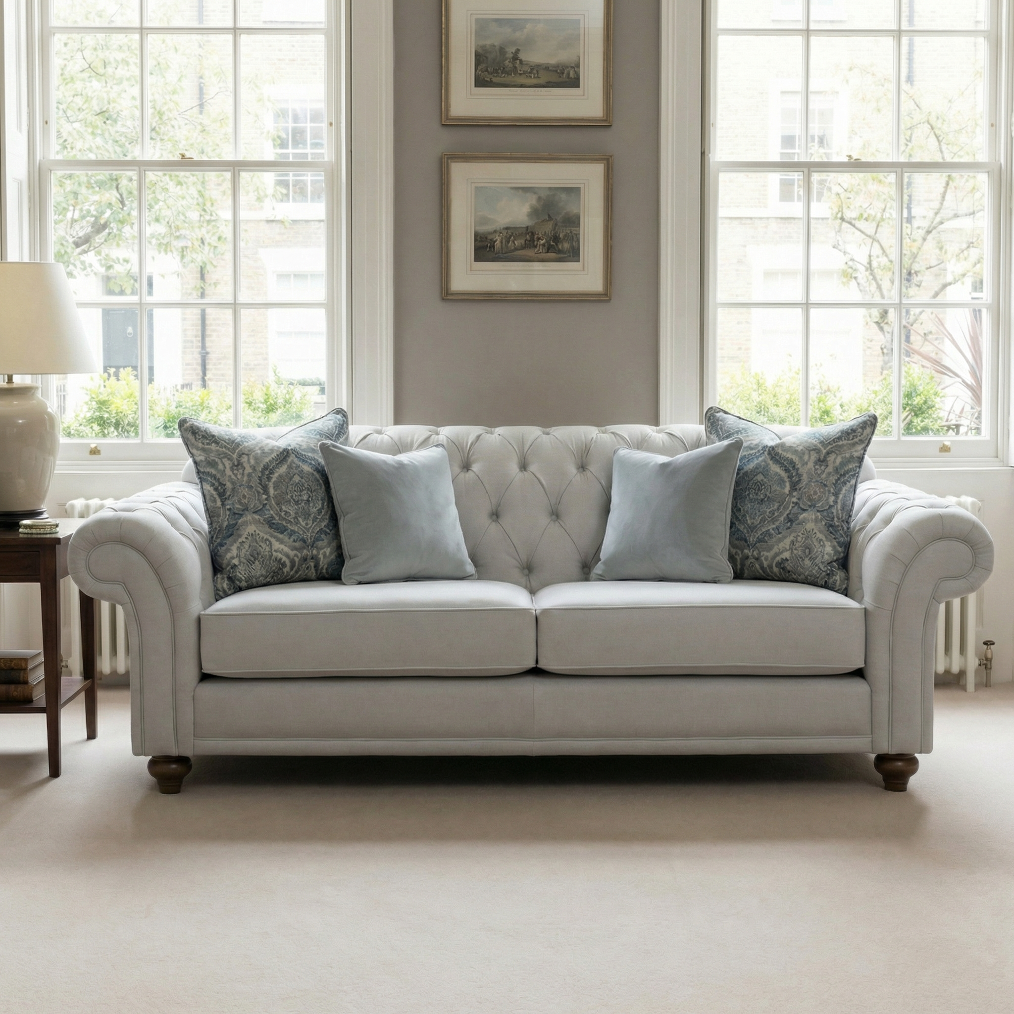 A Chesterfield 3 Seat Sofa in Warm Grey with hand-finished details and patterned and plain pillows is placed in front of two large windows.