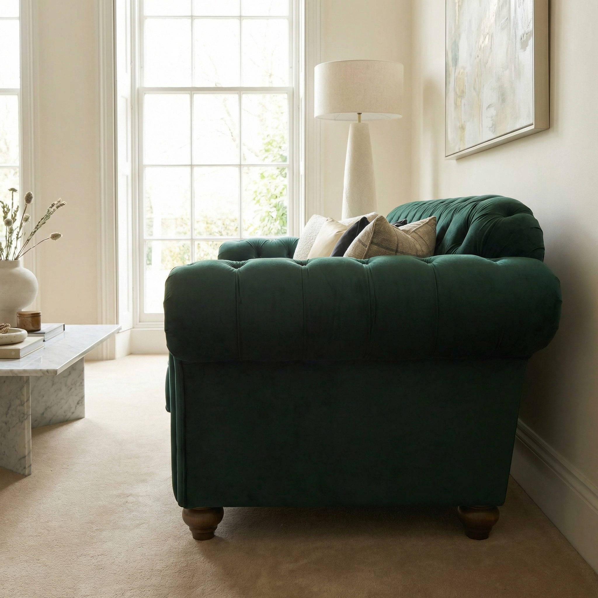 A Chesterfield 3 Seat Velvet Sofa in Bottle Green with hand-finished upholstery is placed beside a window, lamp, and marble coffee table in a bright room.