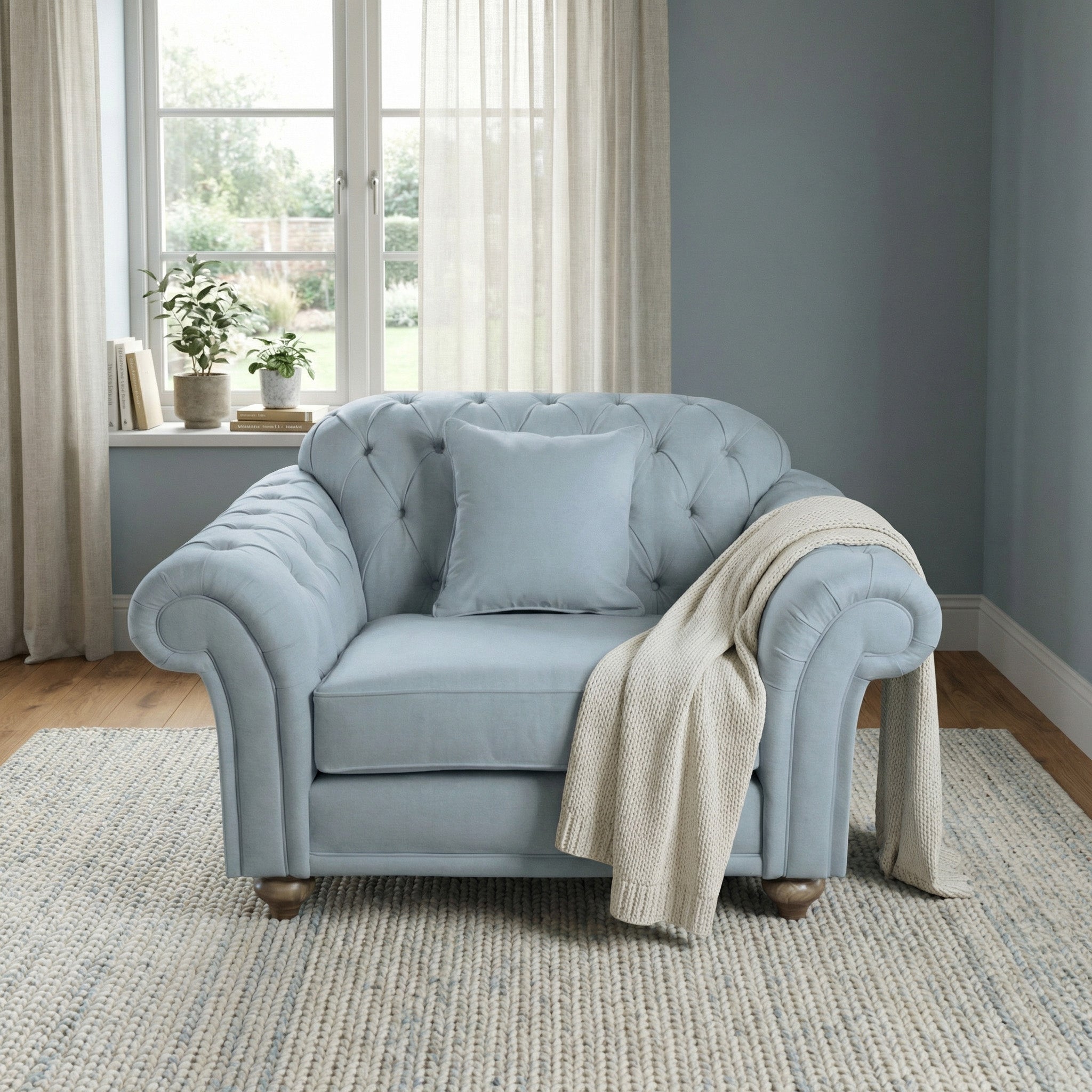 The Chesterfield Loveseat in Sky Blue, paired with a cushion and cream throw on a rug by the window with sheer curtains, adds handcrafted comfort and style to any room.