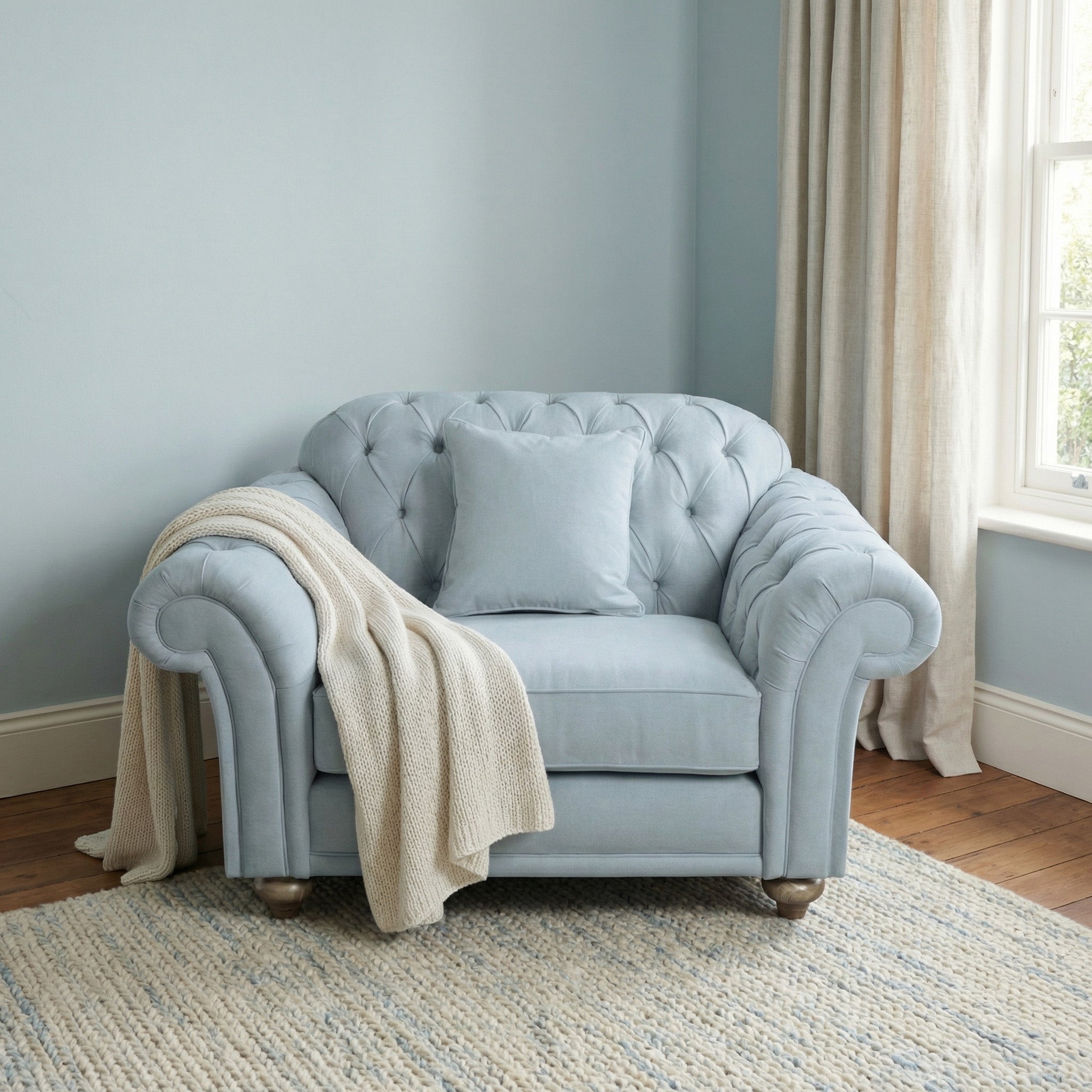The Chesterfield Loveseat in Sky Blue, topped with a cushion and cream knit throw, sits by a window with beige curtains on a rug.