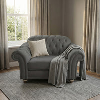 The Chesterfield Loveseat in Smoke Grey, handmade in the UK for timeless elegance and comfort, sits on a rug in a softly lit room with large windows and curtains, styled with a pillow and blanket.