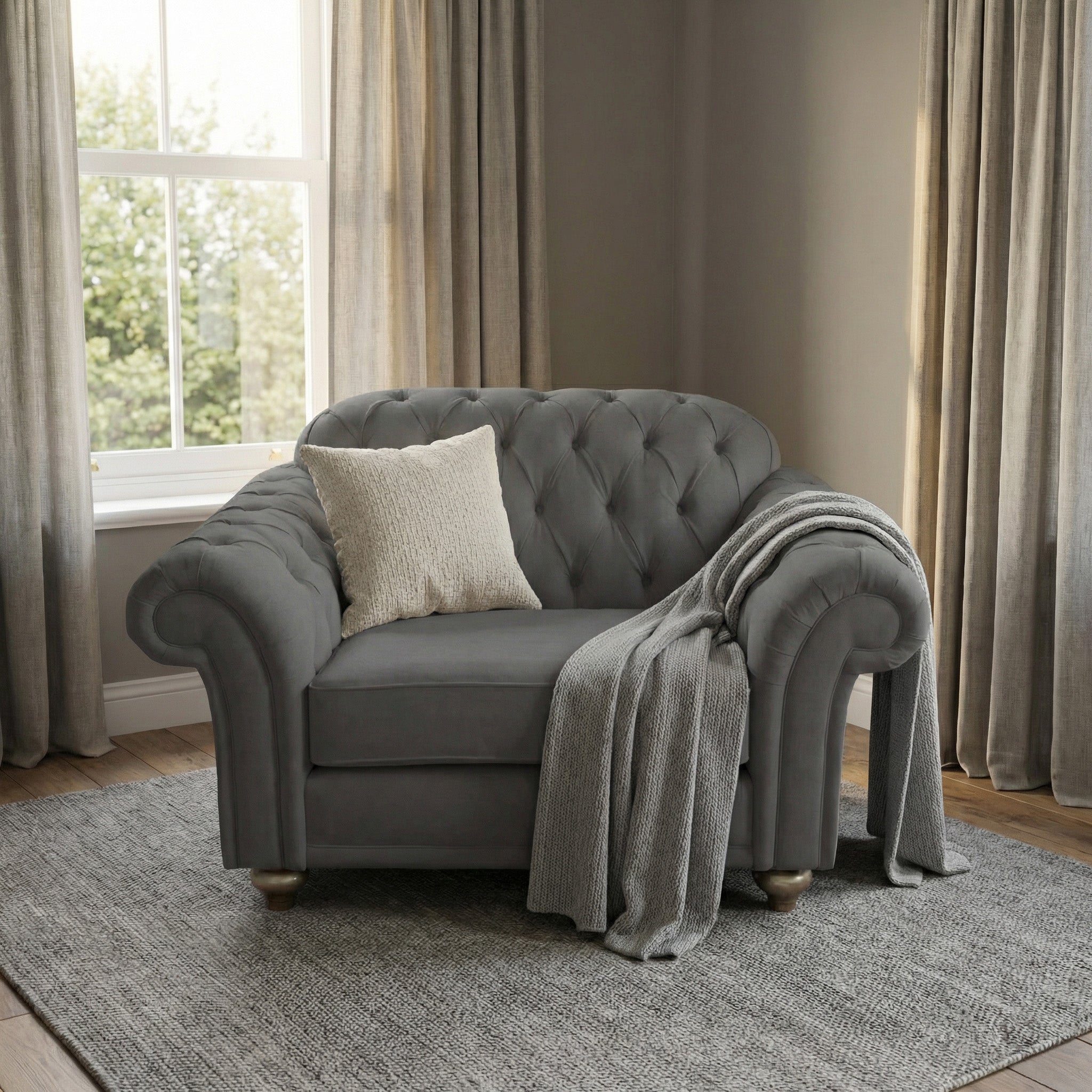 The Chesterfield Loveseat in Smoke Grey, handmade in the UK for timeless elegance and comfort, sits on a rug in a softly lit room with large windows and curtains, styled with a pillow and blanket.