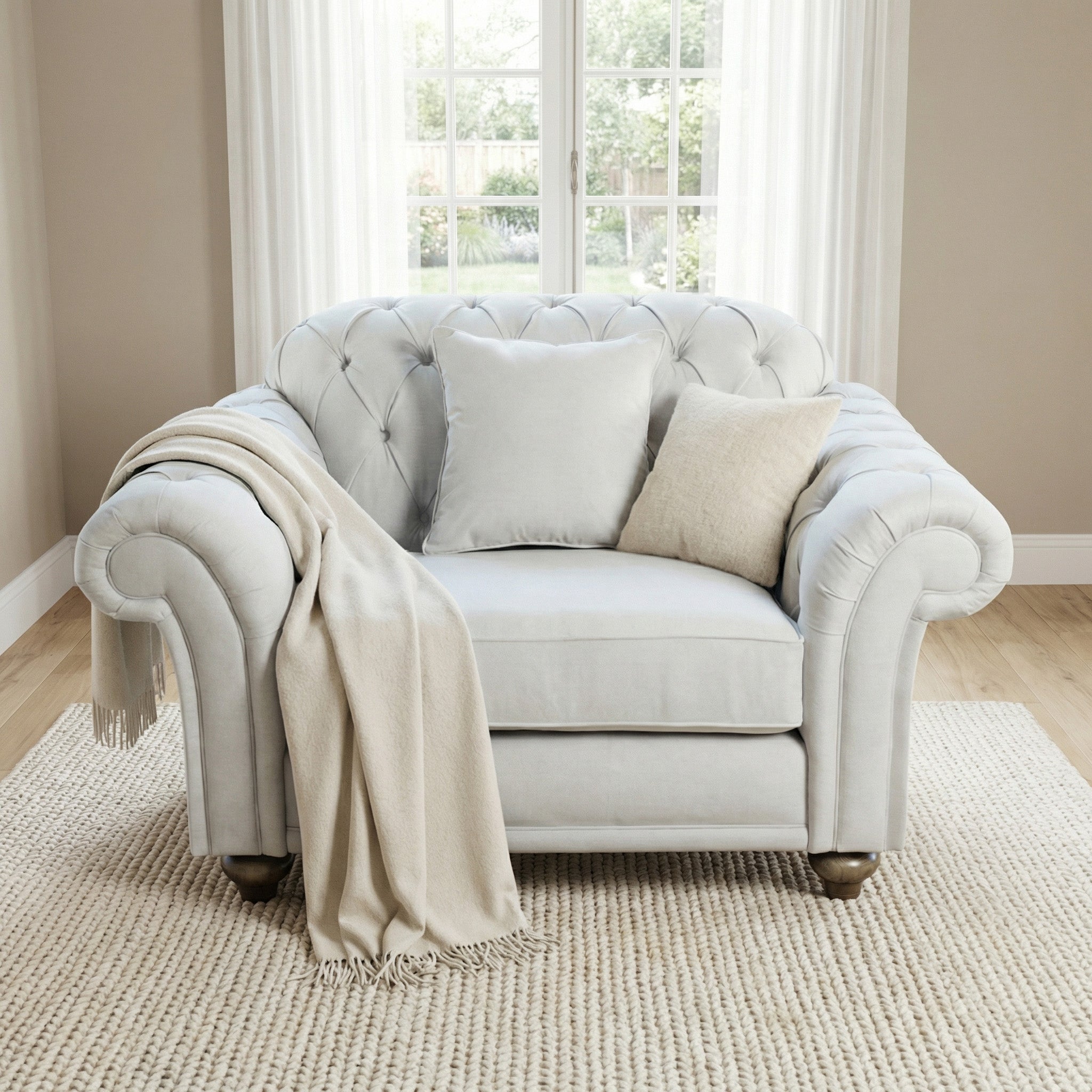 The Chesterfield Loveseat in Warm Grey features tufted upholstery, two pillows, and a beige throw blanket. Placed on a rug by the window, it's crafted from sustainable wood for an eco-friendly touch.