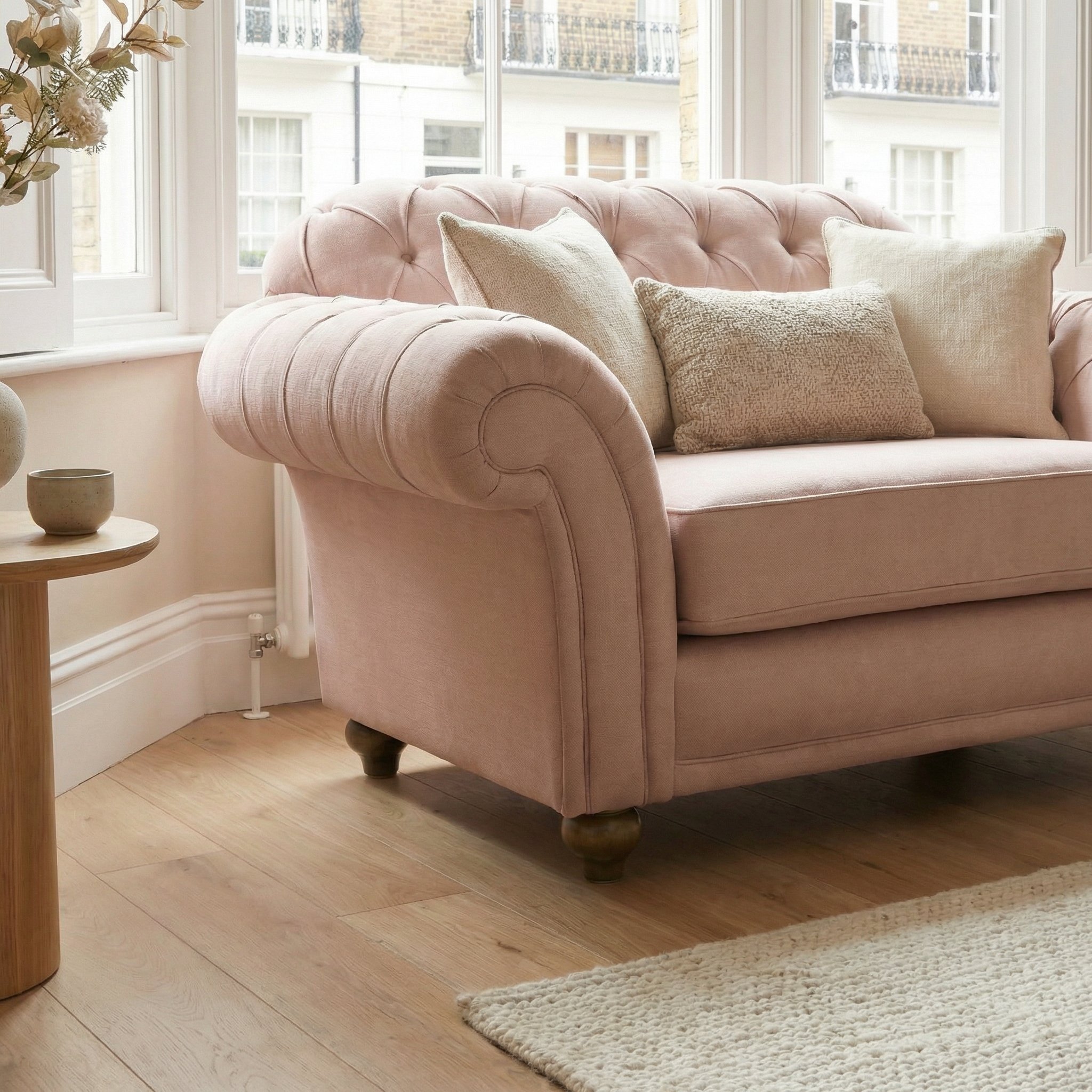 Chesterfield Loveseat in Dusky Pink with beige pillows, set in a bright room with wooden floors and large windows, handcrafted in England.