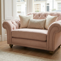 The Chesterfield Loveseat in Dusky Pink, handcrafted in England and paired with three beige cushions, sits by a window in a bright living room.