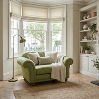 The Chesterfield Loveseat in Olive, complete with plush cushions and a throw, sits by bay windows in a bright, stylish living room.