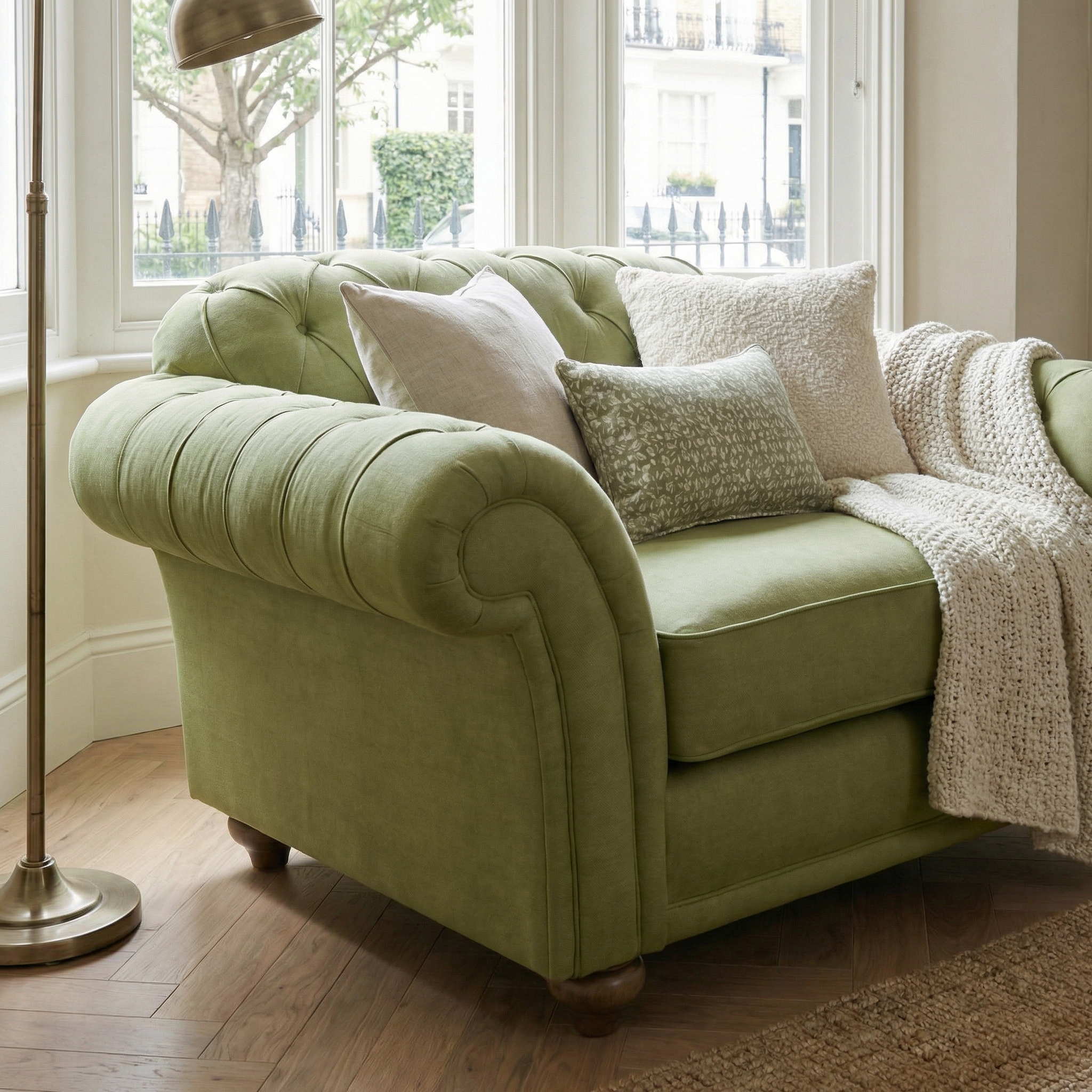 Chesterfield Loveseat in Olive with cushions and a throw, placed by a window and brass floor lamp on wooden flooring.