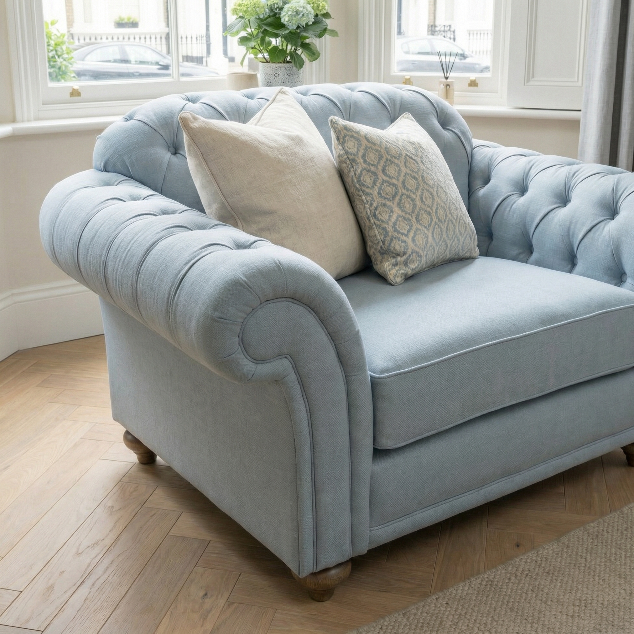 Sky blue Chesterfield Loveseat styled with two patterned cushions in a bright room with bay windows, paired with a handcrafted sofa for sustainable elegance.