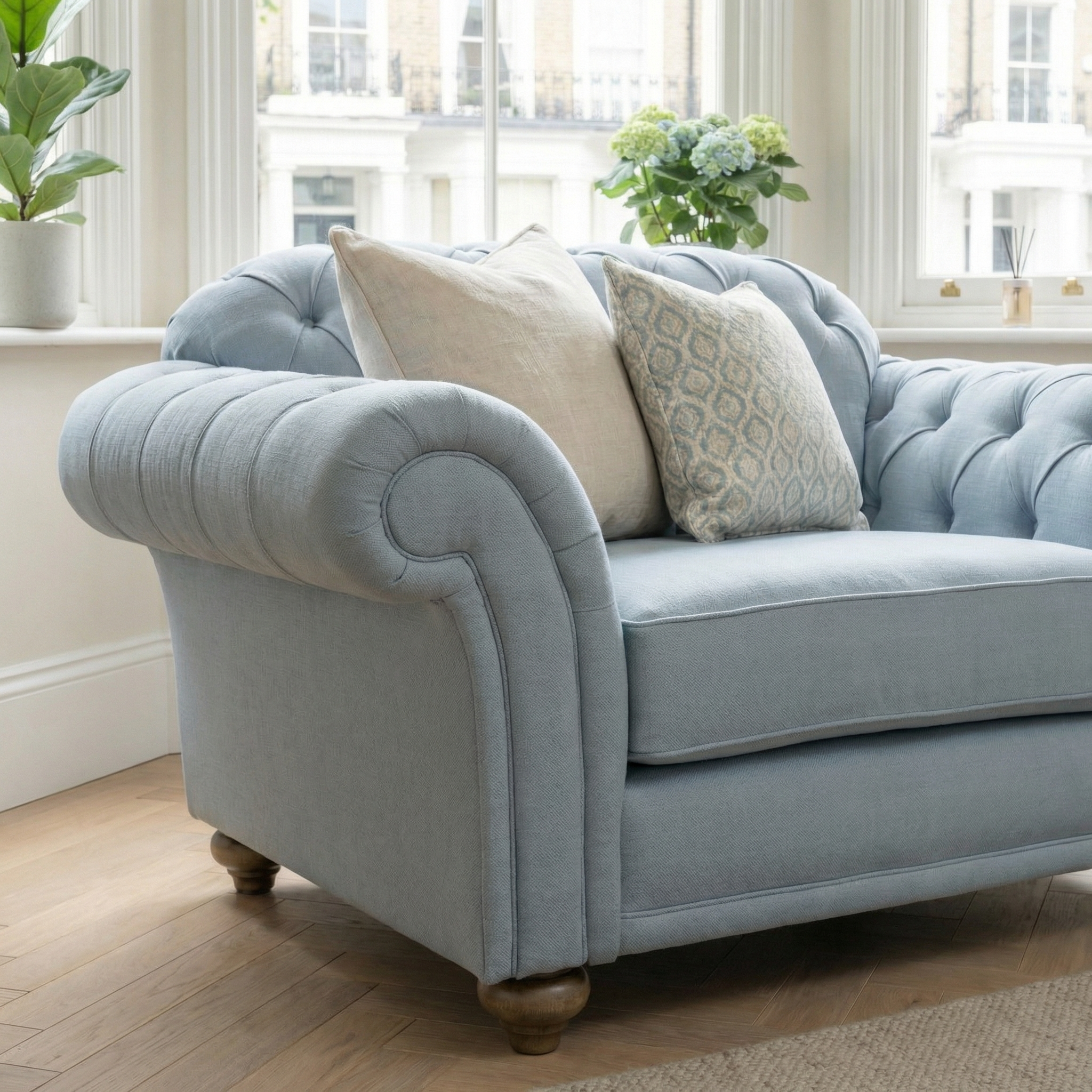 A Chesterfield Loveseat in Sky Blue sits by a window with hydrangeas and a plant, embodying sustainable elegance. Two decorative pillows complete the serene setting.