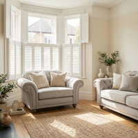 Bright living room featuring a Chesterfield Loveseat in Warm Grey, cozy cushions, a jute rug, lush plants, and large bay windows with white shutters.