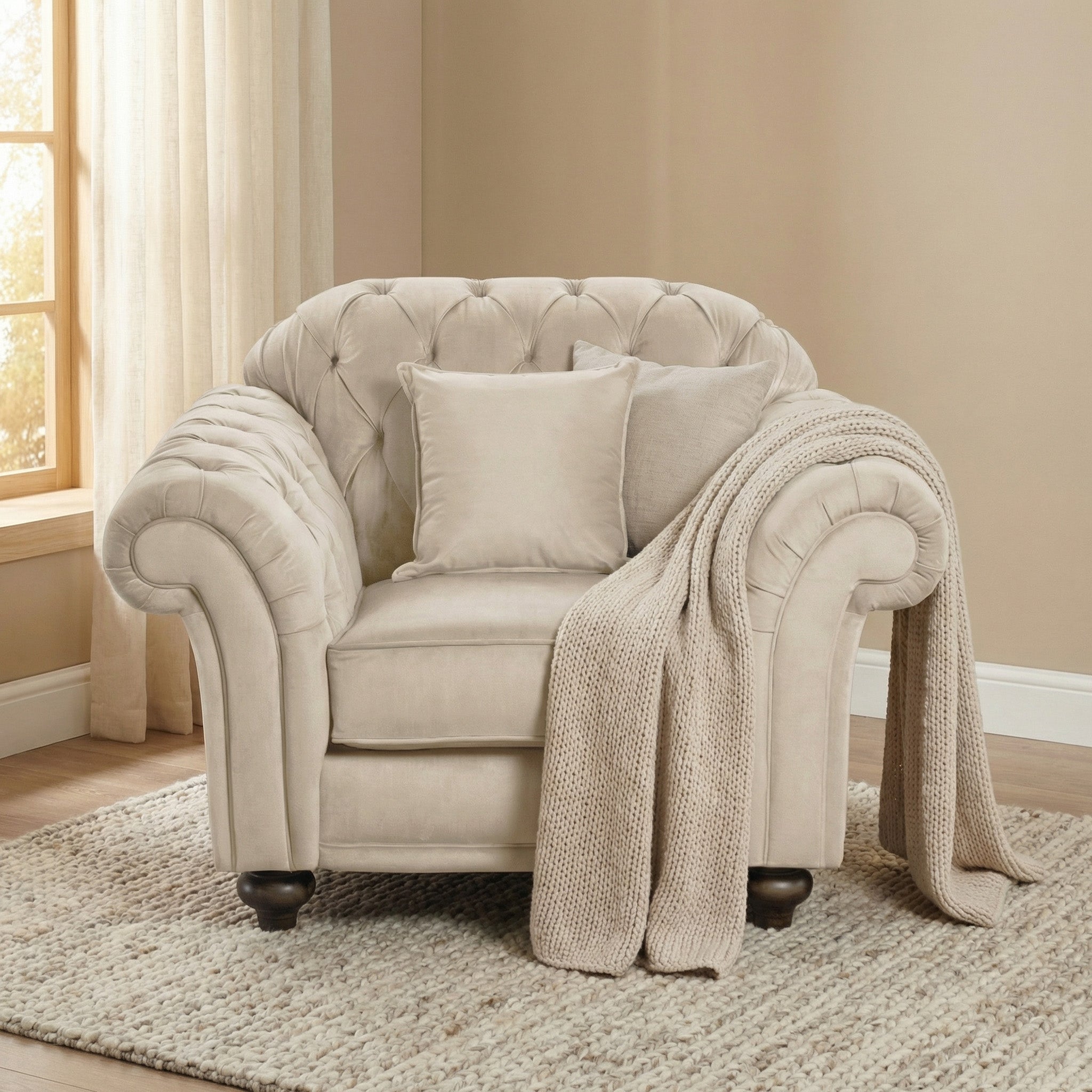 The Chesterfield Velvet Armchair in Cream, featuring plush cushions and a knit throw, sits on a rug near a window with sheer curtains.