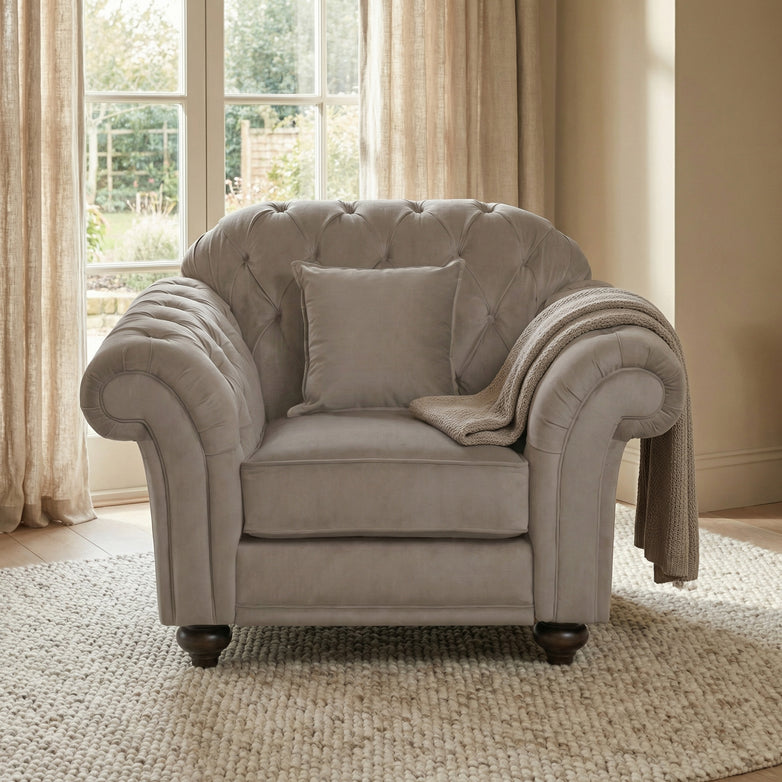 The Chesterfield Velvet Armchair in Mink with a cushion and throw blanket sits on a textured rug in front of large windows.