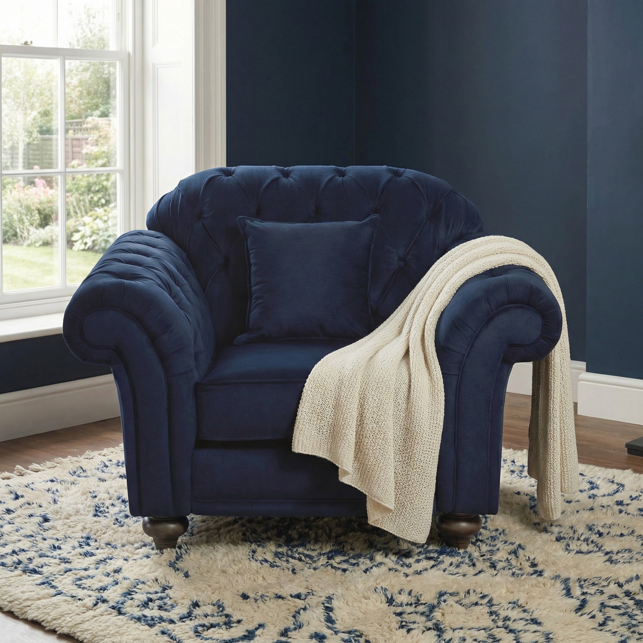 A Chesterfield Velvet Armchair in Royal Blue with a pillow and cream knit throw is placed on a patterned rug by the window.
