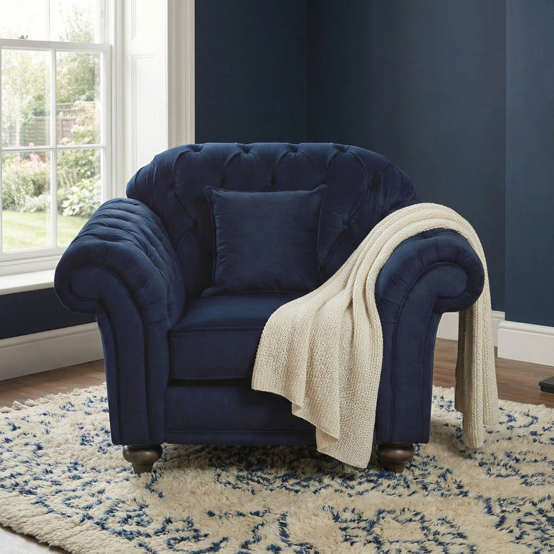 A Chesterfield Velvet Armchair in Royal Blue with a pillow and cream knit throw is placed on a patterned rug by the window.