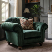 The Chesterfield Velvet Armchair in Bottle Green with a patterned cushion sits in a cozy, elegant living room with gray walls and a plant.