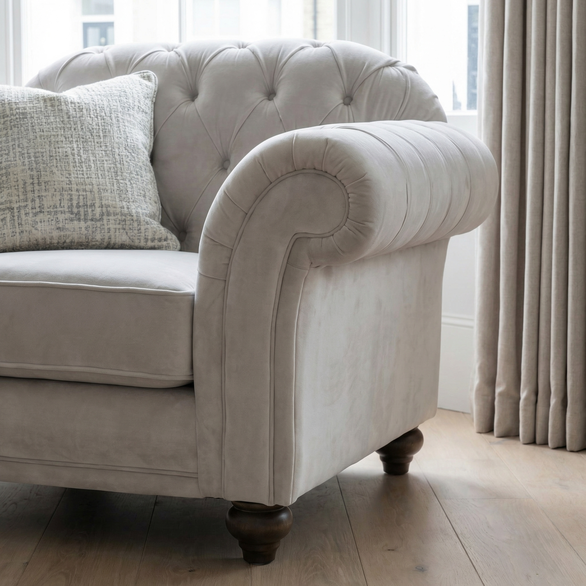 The Chesterfield Velvet Armchair in Feather Grey, featuring elegant tufting and a patterned cushion, sits on a wooden floor near a window with beige curtains.