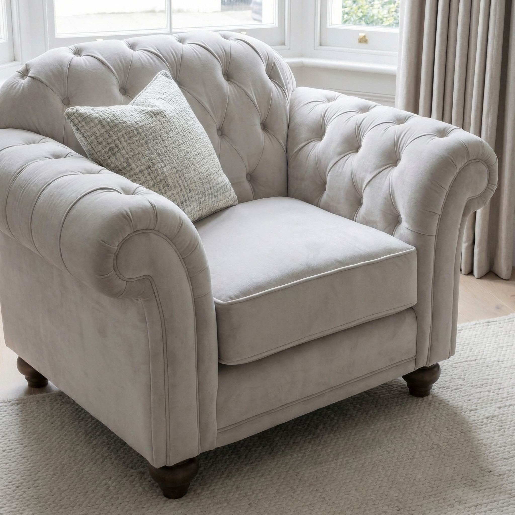 A Chesterfield Velvet Armchair in Feather Grey with a patterned cushion sits by a window framed with beige curtains.